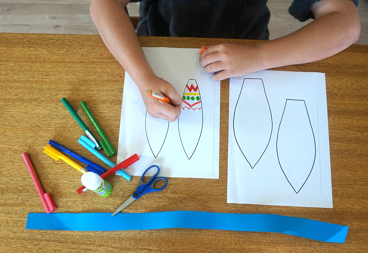 Boy colouring in bunny ear with textas while the rest of the required equipment sits on table including Paper Templates, Scissors, Glue Stick, Paper Ribbon, Coloured Textas and a Stapler