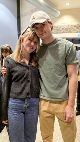 Milla and Rowan A young couple. She has blonde long hair wearing a grey jacket and jeans. He has his arm around her and is wearing a cap, tshirt and tan pants