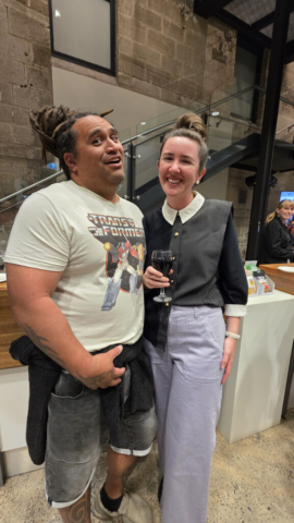 Quentin Wiperi and Belinda Anderson A couple posing for the camera. He has dreaded hair in a bun wearing a transformers tshirt and denim jeans, she has long straight hair in a bun, collared shirt and jeans, and is holding a glass of wine.