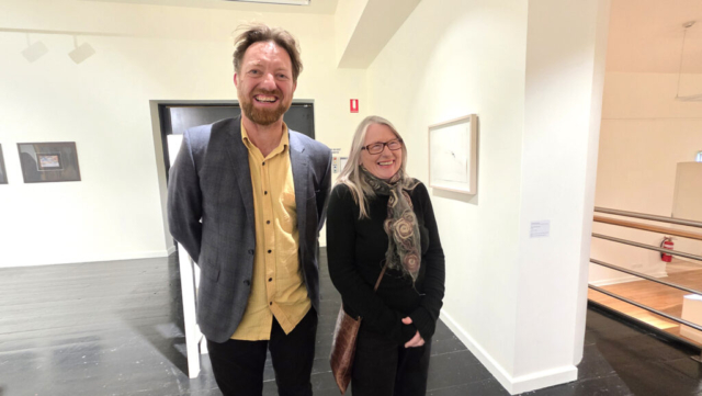 Curator Chris Clements, and artist Carmel Wallace A tall smiling man with a grey jacket and yellow shirt next to a smiling woman with long straight hair and glasses