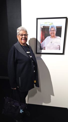 Aunty Val Brennan with her portrait An Aboriginal elder with short grey hair is wearing a navy jacket and stands next to her portrait.