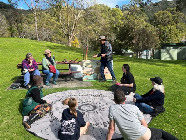 Walking the seasons An Aboriginal Elder speaks to a group of people sitting around a rug. They are on lush green grass with trees in the background.